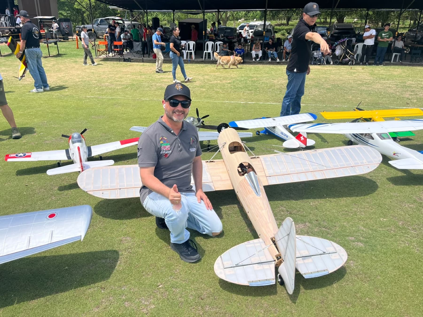 Un socio del club agachado junto a un avión de madera sin entelar, con otros modelos y pilotos detrás.