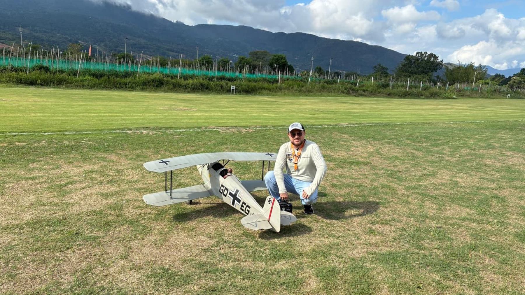 Piloto agachado junto a un biplano a escala de época, blanco con cruces negras, en medio de la pista de pasto con montañas al fondo.