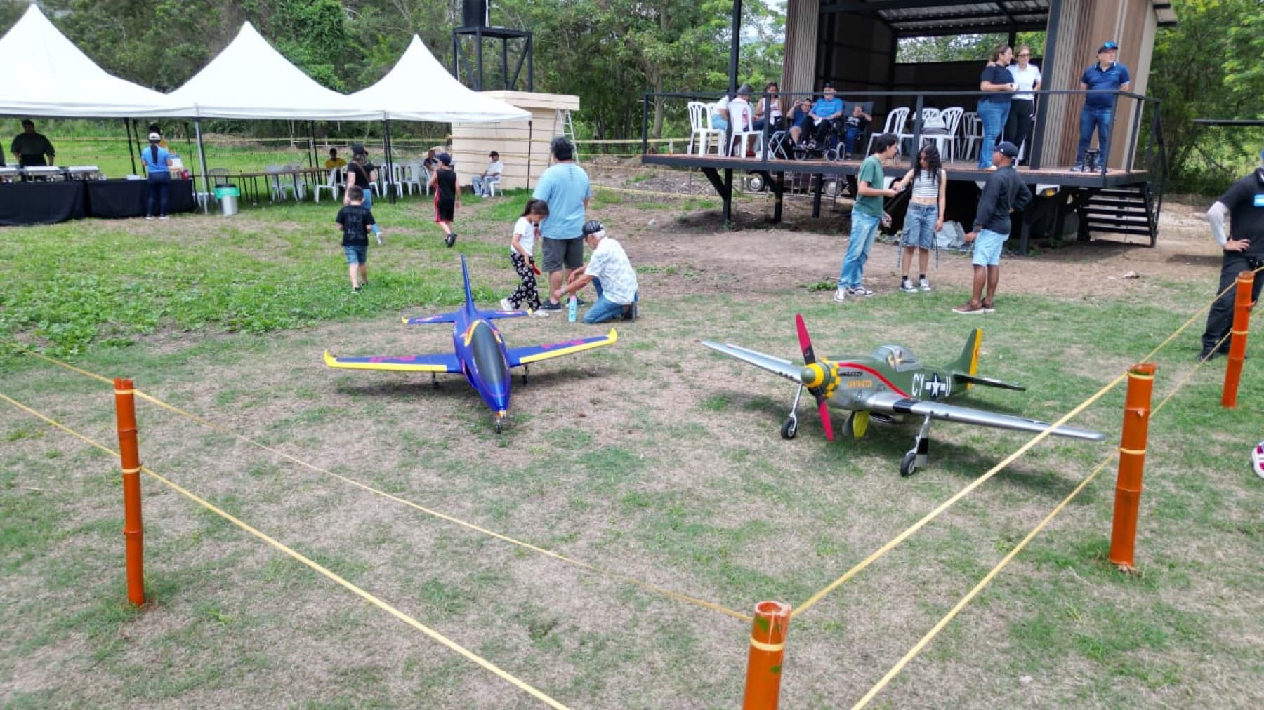 Un jet azul y un P-51 Mustang sobre el césped dentro de una zona acordonada, con público y carpas al fondo.