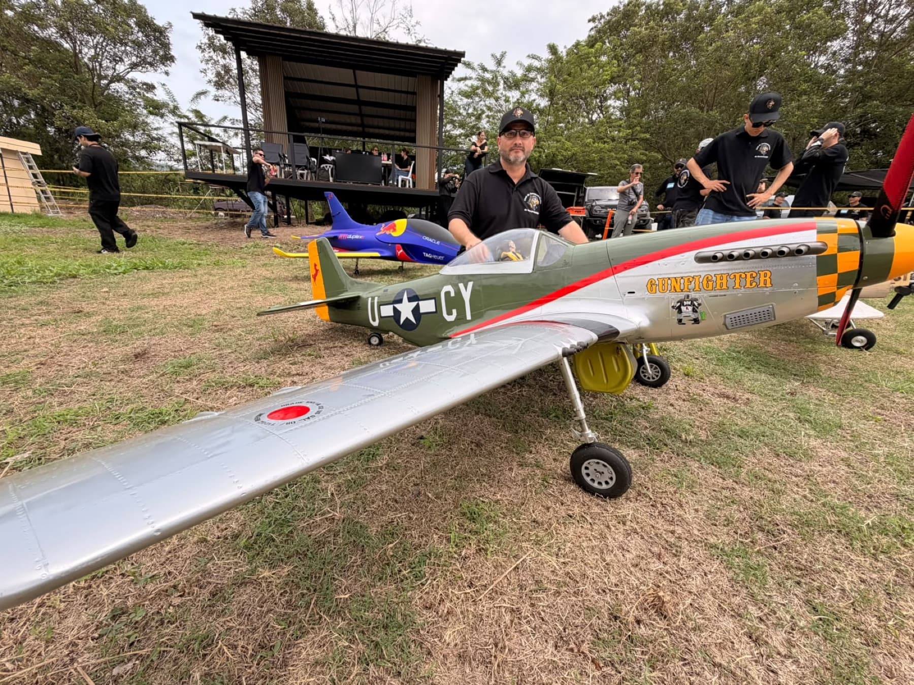Piloto de pie detrás de un P-51 Mustang a escala grande con nariz amarilla y franjas de invasión, sobre el pasto del campo.