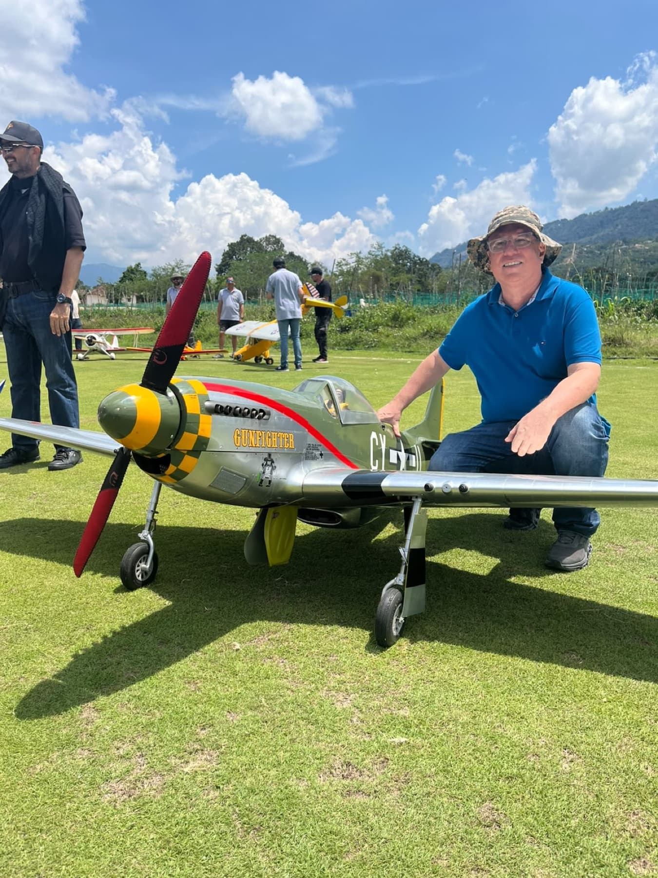 Piloto agachado junto a un P-51 Mustang a escala, con hélice de cuatro palas, tren principal desplegado y pintura plateada de la Segunda Guerra Mundial.