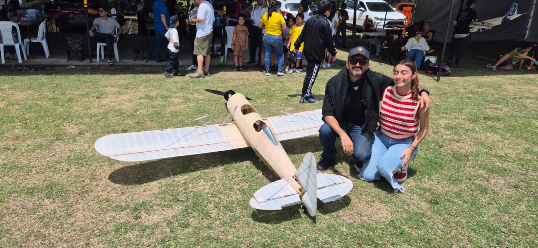 Un piloto del club y su hija sonríen junto a un aeromodelo de madera a escala sobre el césped.