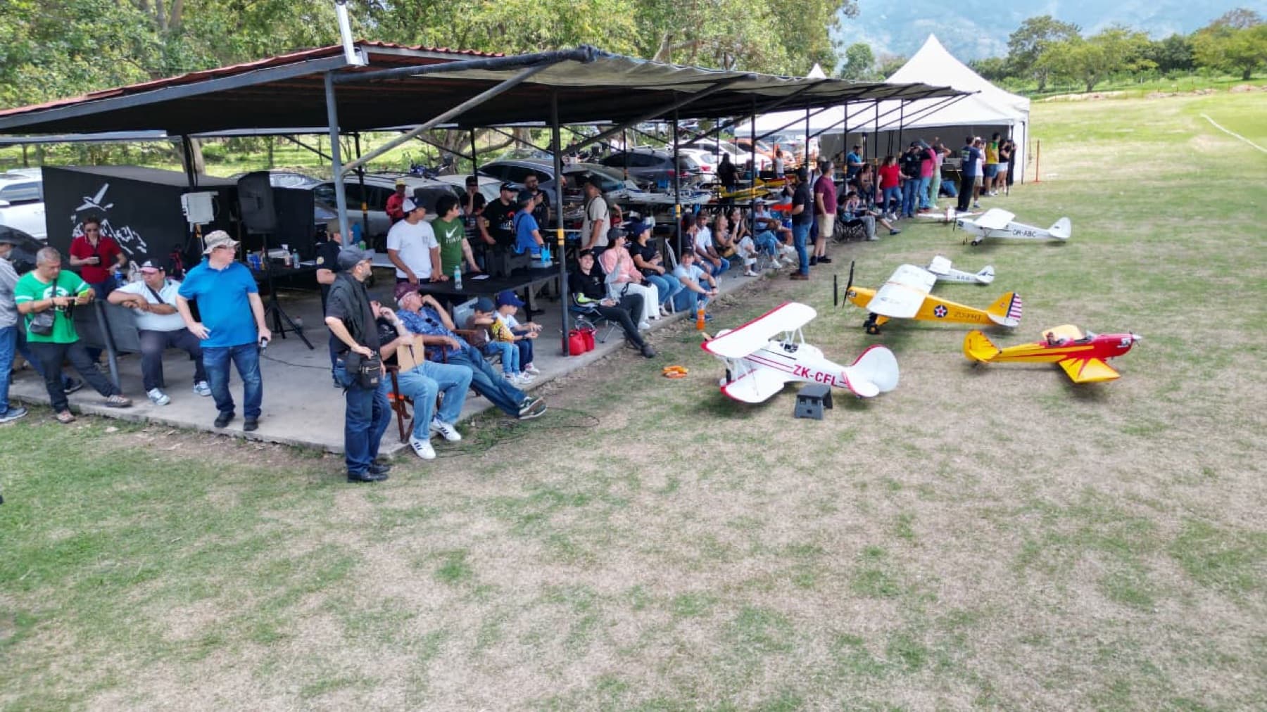 Vista aérea de los pits del club: un techo con pilotos y público a la sombra y varios aviones sobre el césped.